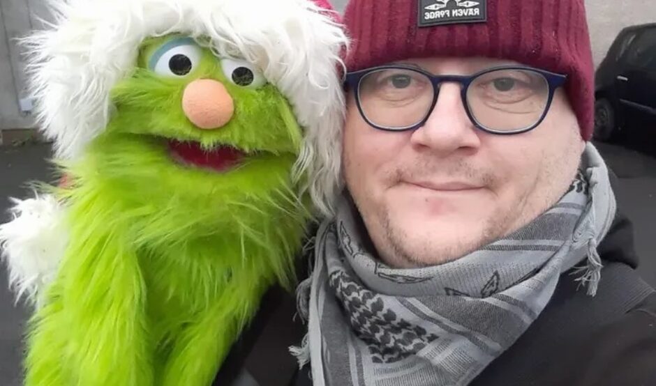 A selfie of a man wearing a maroon beanie and glasses, smiling next to a green Kermit-like puppet wearing a Santa hat.