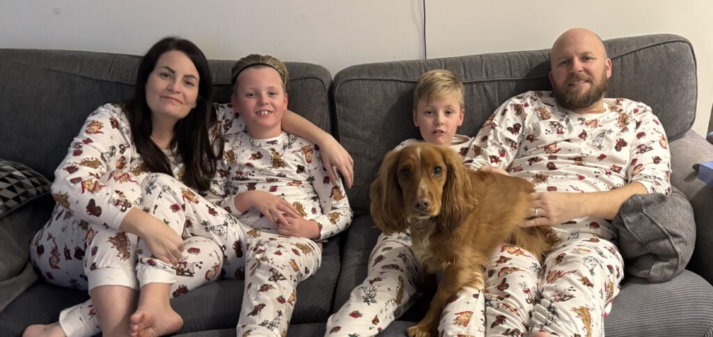 A family of four: Sophie, a man with a beard, and two young boys, sit together on a grey sofa smiling, all wearing matching white holiday pyjamas with festive patterns. A medium-sized golden-brown dog sits comfortably across the laps of the man and one of the boys.