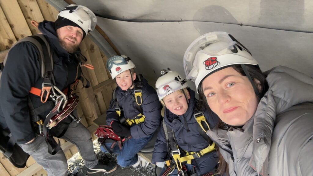 A family selfie of Sophie, her husband, and their twin boys geared up for ziplining at Zip World. Everyone is wearing safety harnesses and white helmets with the "Zip World" logo, posing together inside a covered wooden staging or waiting area.