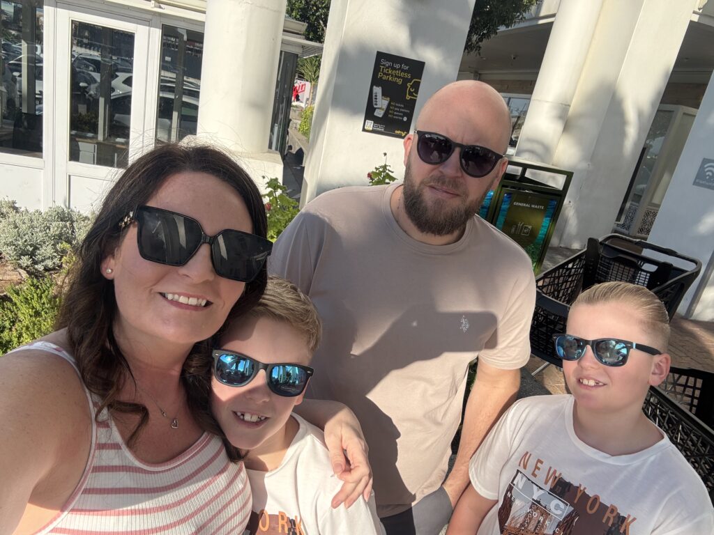 An outdoor family selfie of Sophie, her husband, and their twin boys on a sunny day. Sophie, on the left, smiles while wearing large black sunglasses and a striped tank top. Her husband, a bald man with a beard in a tan t-shirt, stands behind them, and both of their twin boys smile while wearing matching sunglasses with reflective blue lenses.