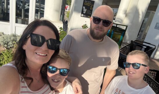 An outdoor family selfie of Sophie, her husband, and their twin boys on a sunny day. Sophie, on the left, smiles while wearing large black sunglasses and a striped tank top. Her husband, a bald man with a beard in a tan t-shirt, stands behind them, and both of their twin boys smile while wearing matching sunglasses with reflective blue lenses.