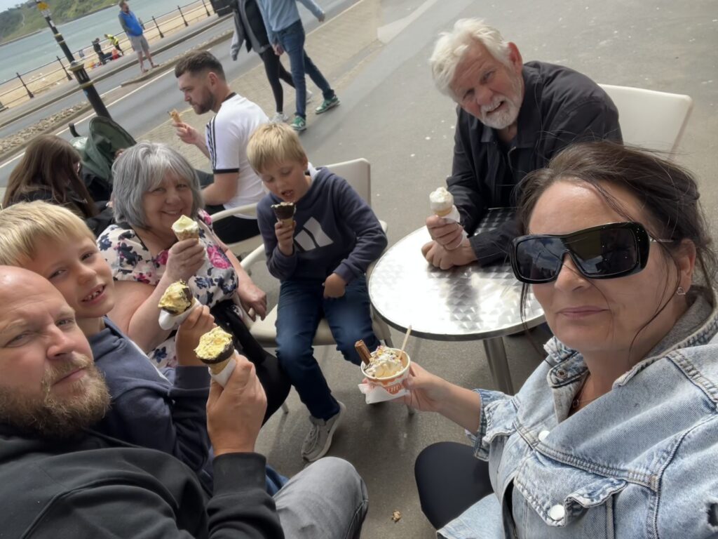 A high-angle family selfie taken outdoors by Sophie, showing a group of six people sitting around an outdoor table enjoying ice cream treats. Sophie is in the foreground wearing large sunglasses and a denim jacket, holding up a small tub of ice cream with a chocolate flake.