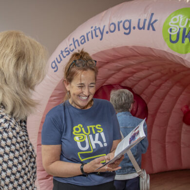 A staff member at Guts UK is wearing a charity t-shirt and holding an information leaflet up whilst talking to a member of the public. Behind them is an inflatable colon.