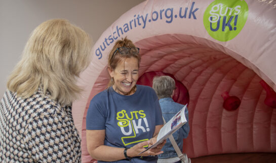 A staff member at Guts UK is wearing a charity t-shirt and holding an information leaflet up whilst talking to a member of the public. Behind them is an inflatable colon.