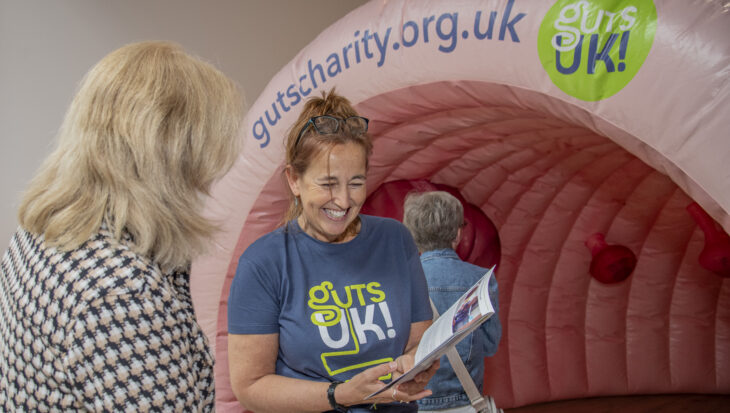 A staff member at Guts UK is wearing a charity t-shirt and holding an information leaflet up whilst talking to a member of the public. Behind them is an inflatable colon.