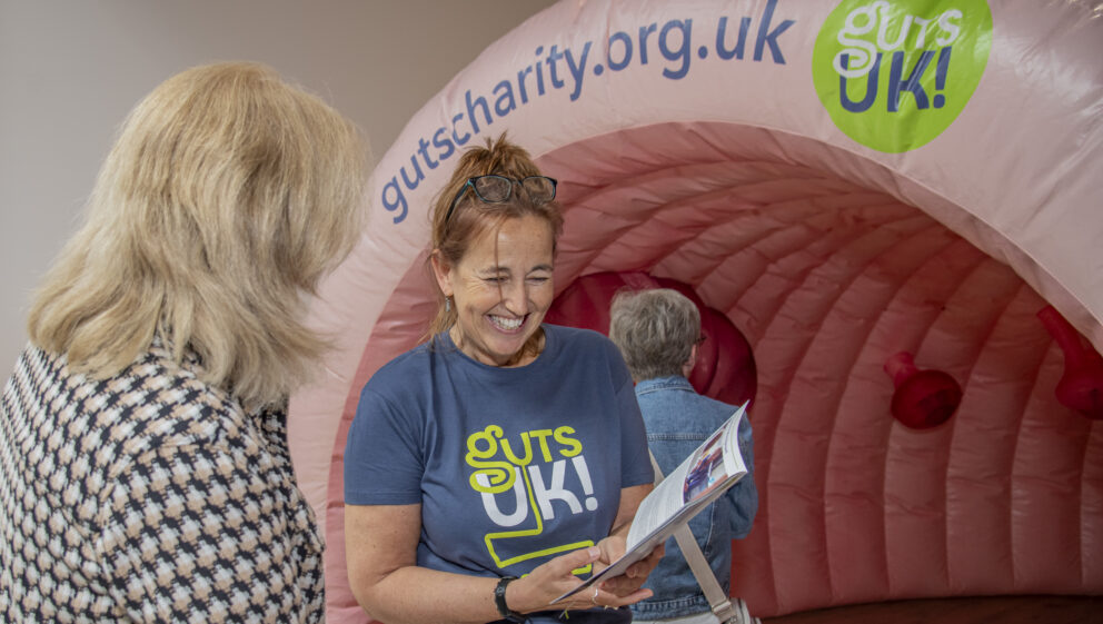 A staff member at Guts UK is wearing a charity t-shirt and holding an information leaflet up whilst talking to a member of the public. Behind them is an inflatable colon.