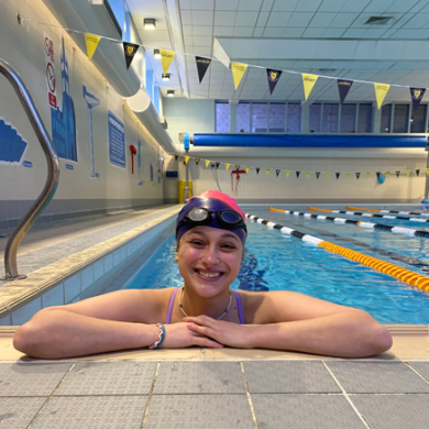 A close-up, eye-level shot of a smiling woman resting her arms on the tiled edge of an indoor swimming pool. She is wearing a pink and black swim cap, dark goggles resting on her forehead, a purple swimsuit strap, and a thin beaded necklace and bracelet. The background shows the clear blue water of the pool lanes divided by orange and black floats, under an indoor ceiling with hanging yellow and dark pennant banners.