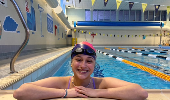 A close-up, eye-level shot of a smiling woman resting her arms on the tiled edge of an indoor swimming pool. She is wearing a pink and black swim cap, dark goggles resting on her forehead, a purple swimsuit strap, and a thin beaded necklace and bracelet. The background shows the clear blue water of the pool lanes divided by orange and black floats, under an indoor ceiling with hanging yellow and dark pennant banners.