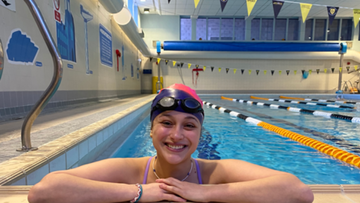 A close-up, eye-level shot of a smiling woman resting her arms on the tiled edge of an indoor swimming pool. She is wearing a pink and black swim cap, dark goggles resting on her forehead, a purple swimsuit strap, and a thin beaded necklace and bracelet. The background shows the clear blue water of the pool lanes divided by orange and black floats, under an indoor ceiling with hanging yellow and dark pennant banners.
