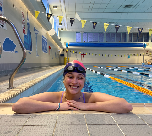 A close-up, eye-level shot of a smiling woman resting her arms on the tiled edge of an indoor swimming pool. She is wearing a pink and black swim cap, dark goggles resting on her forehead, a purple swimsuit strap, and a thin beaded necklace and bracelet. The background shows the clear blue water of the pool lanes divided by orange and black floats, under an indoor ceiling with hanging yellow and dark pennant banners.