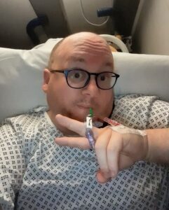 A selfie of a man wearing a hospital gown and glasses, holding up a peace sign with an IV port taped to the back of his hand.