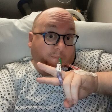 A selfie of a man wearing a hospital gown and glasses, holding up a peace sign with an IV port taped to the back of his hand.