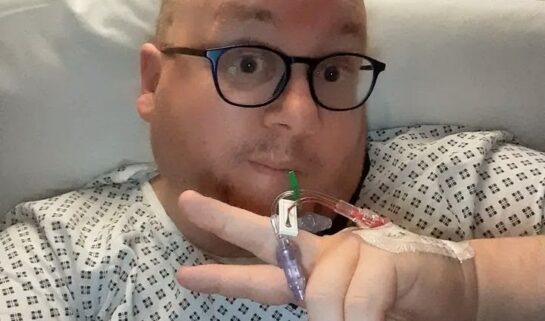 A selfie of a man wearing a hospital gown and glasses, holding up a peace sign with an IV port taped to the back of his hand.