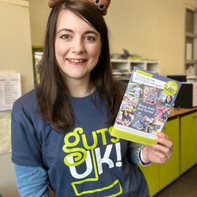 Amy is smiling, wearing a Guts UK t-shirt, poop emoji boppers on her head and holds a Guts UK magazine in her hand.