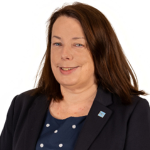 Headshot of Sarah, a woman with shoulder-length brown hair, smiling and wearing a dark blue blazer over a blue polka-dot top, set against a plain white background.