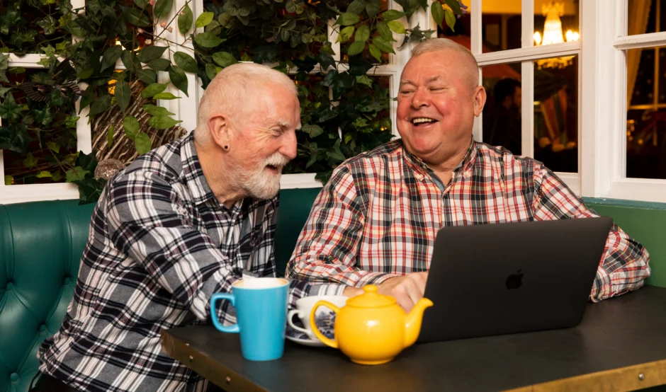 Two men sat next to each other talking at a table. There is a blue mug and a yellow tea pot on the table and they are looking at a laptop. They're both wearing checked shirts and are laughing as they talk.