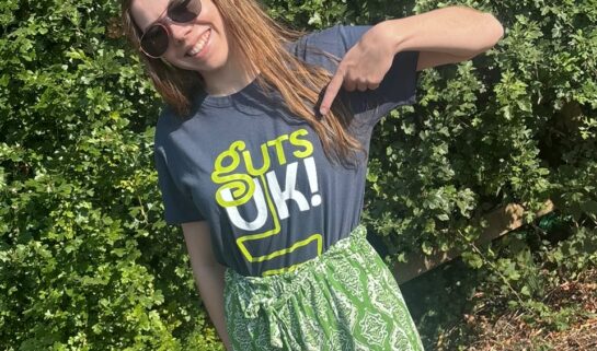 Beth is a young woman standing in a park wearing sunglasses, green shorts and a 'Guts UK' branded top and she is pointing to the logo on the top
