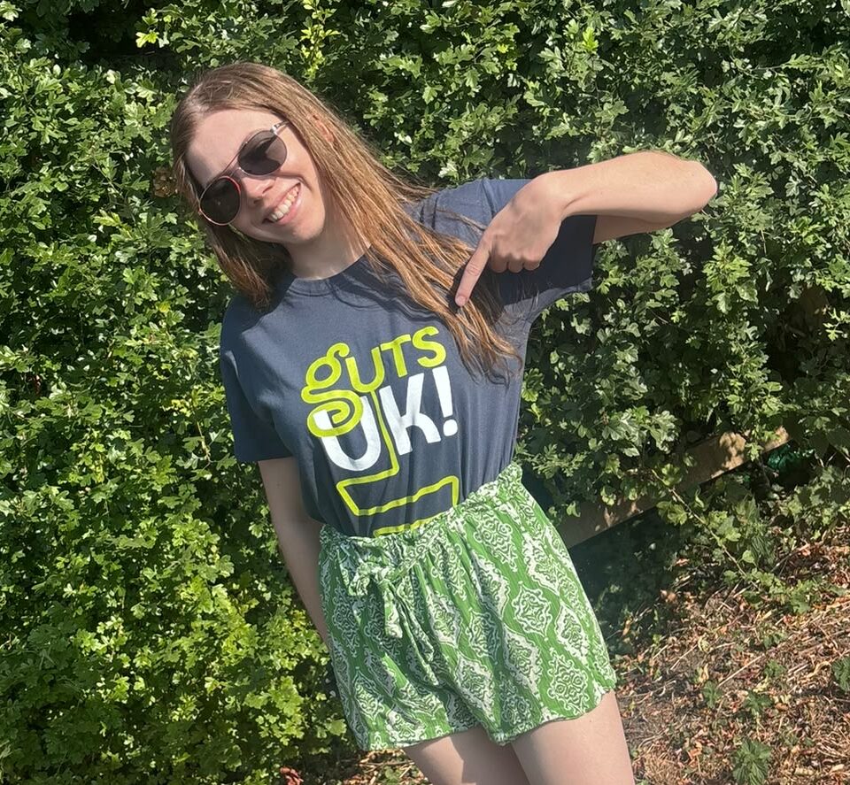 Beth is a young woman standing in a park wearing sunglasses, green shorts and a 'Guts UK' branded top and she is pointing to the logo on the top