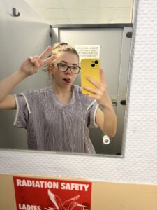 Beth is a young woman in a hospital robe in a hospital setting looking at the mirror doing a selfie playfully sticking her tongue out while doing a peace sign