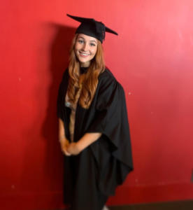 AR is smiling and posing in front of a red wall, wearing a black graduation gown and hat.