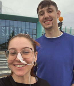 A selfie of AR wearing glasses and a nasal tube, smiling alongside Callum outdoors in front of a building with glass windows.