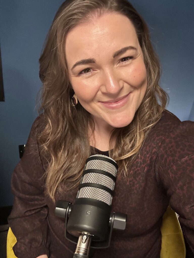 A selfie of Rachel smiling directly into the camera while sitting in a studio setup. In the foreground is a professional silver and black studio microphone. She is wearing a dark patterned long-sleeve top and gold hoop earrings against a dark blue wall.