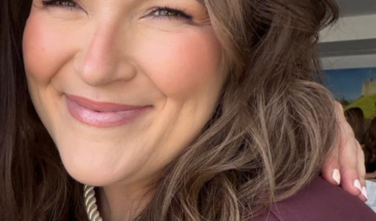 Close-up selfie of Rachel smiling with soft, wavy brown hair framed around her face. She is wearing a maroon top, a gold twisted choker necklace, and soft makeup with warm blush and a natural lip colour.