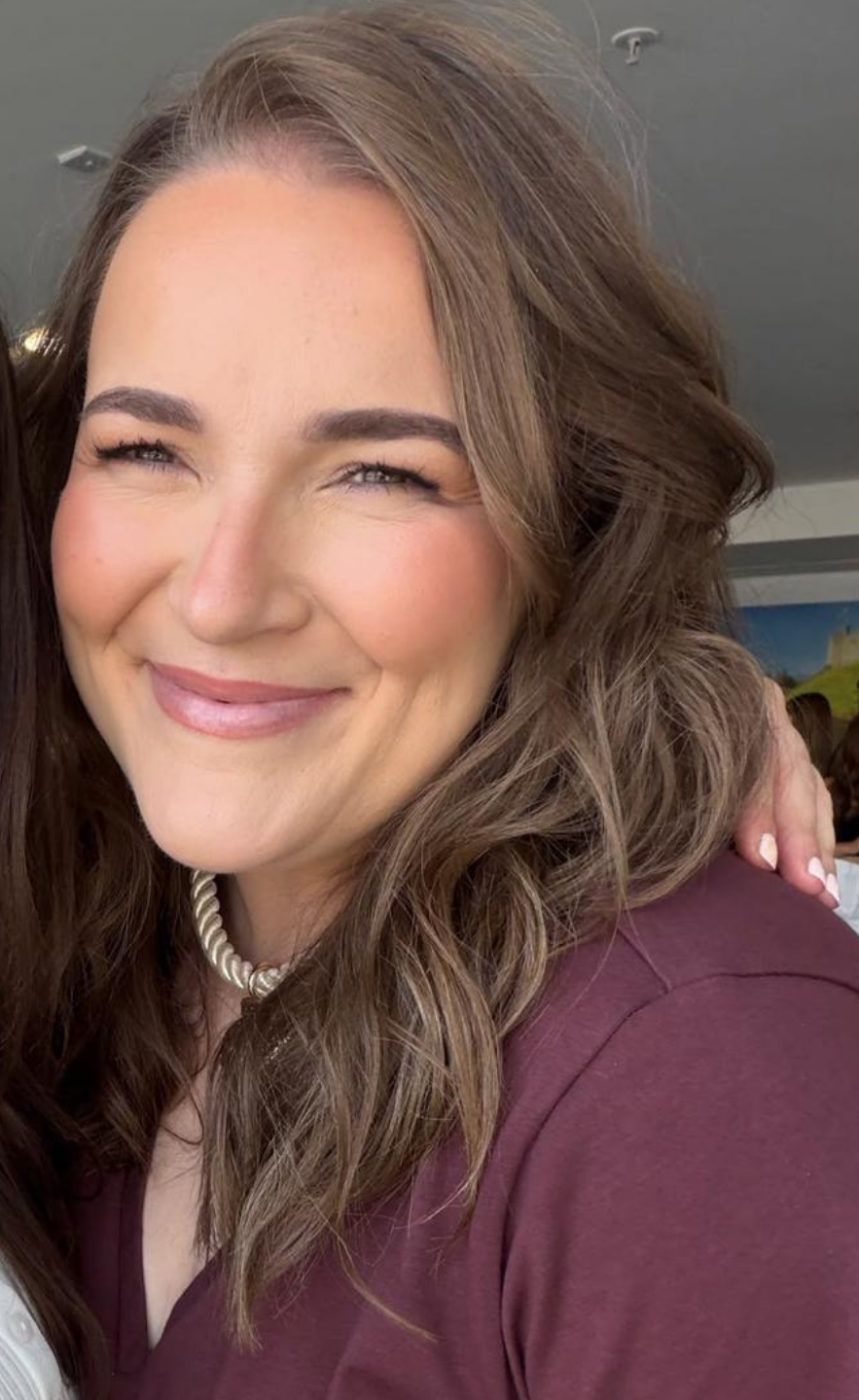 Close-up selfie of Rachel smiling with soft, wavy brown hair framed around her face. She is wearing a maroon top, a gold twisted choker necklace, and soft makeup with warm blush and a natural lip colour.