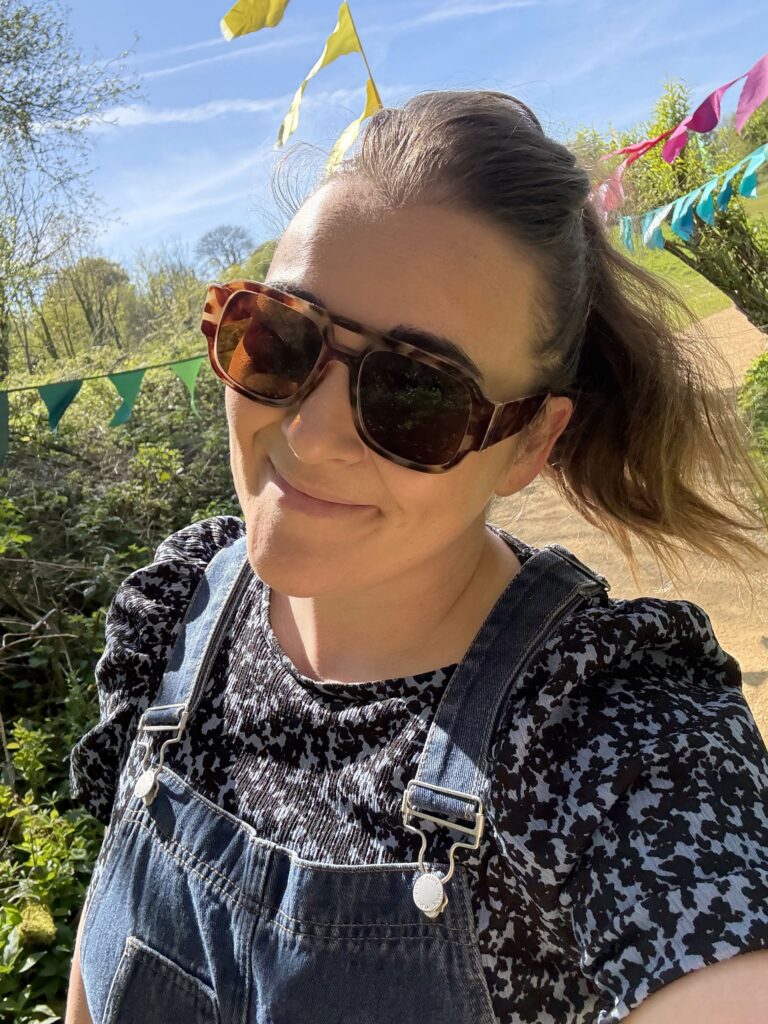 A bright outdoor selfie of Rachel wearing tortoiseshell sunglasses and a casual ponytail. She is wearing denim dungarees over a patterned black-and-white top, with colourful festive bunting overhead under a sunny blue sky.