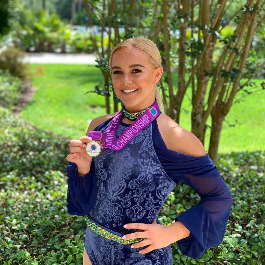 Blonde woman in a blue velvet dance costume smiling outdoors while holding up a medal with a purple championship ribbon.