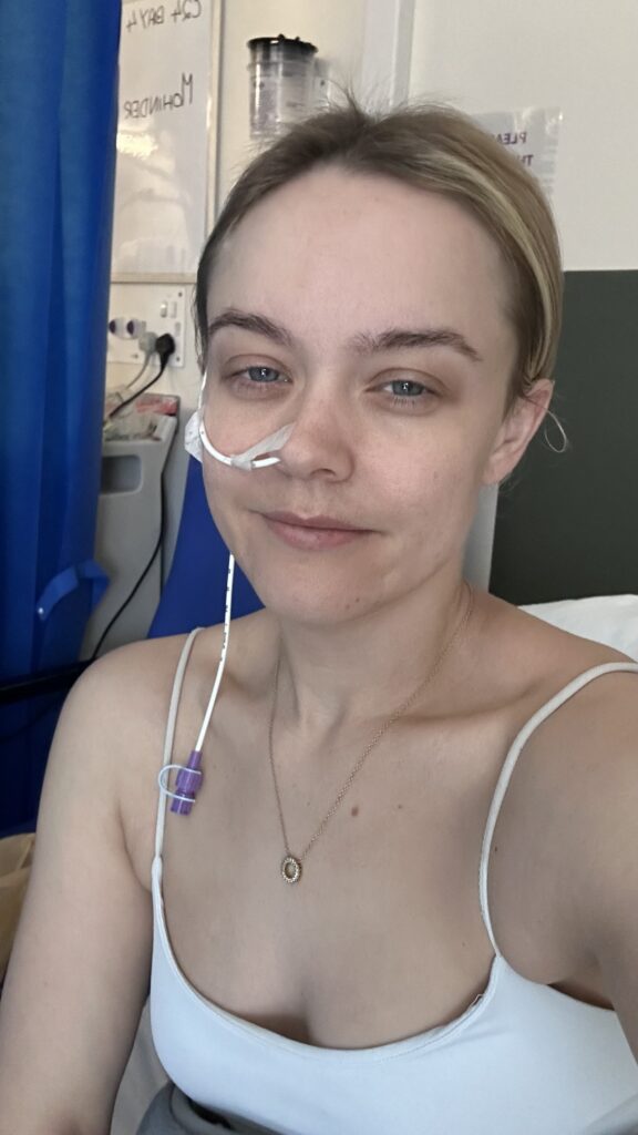 Blonde woman smiling softly in a hospital setting with a clear nasal tube taped to her face.