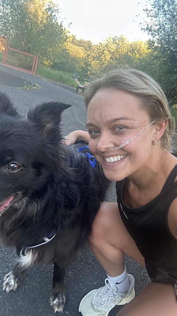Blonde woman with a NG tube smiling outdoors while crouching next to a black border collie.