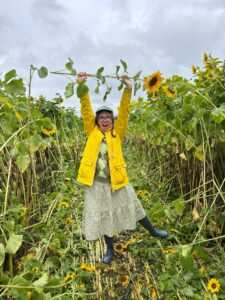 Bella is a young woman standing in a sunflower field playfully holding up a sunflower whilst balancing her leg out to the side and posing playfully
