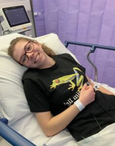 Beth is lying down in a hospital bed smiling to the camera with her thumbs up she is wearing a black t-shirt and is wearing glasses, she has a hospital band around her wrist