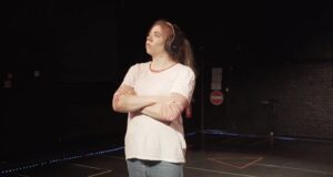 Beth is standing in a theatre practicing her acting for a show the background is dark and she is standing on a stage, wearing headphones with her arms crossed.