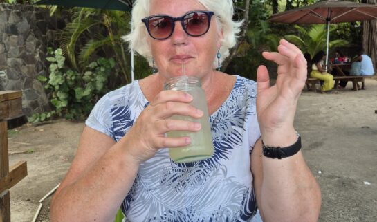Annie is pictured on holiday in St Vincent with trees behind her sitting down, she's wearing a blue top, sunglasses and is drinking a drink