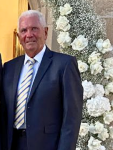 Michael is wearing a dark suit jacket, white shirt, and a blue and yellow striped tie, standing in front of a white floral arrangement.