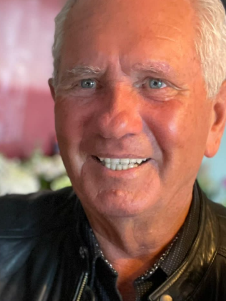 A close-up portrait of Michael smiling with short white hair, light blue eyes, and wearing a black leather jacket.