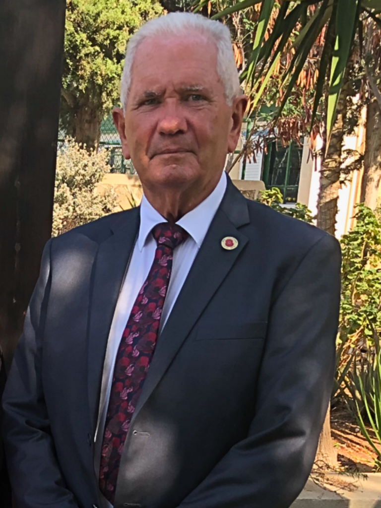 A medium portrait of Michael with short white hair, wearing a dark suit, white shirt, patterned red and purple tie, and a poppy pin on his lapel outdoors.