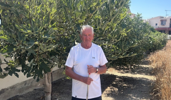 Michael standing outside in Cyprus holding a garden rake in front of tall, leafy fig trees on a sunny day.