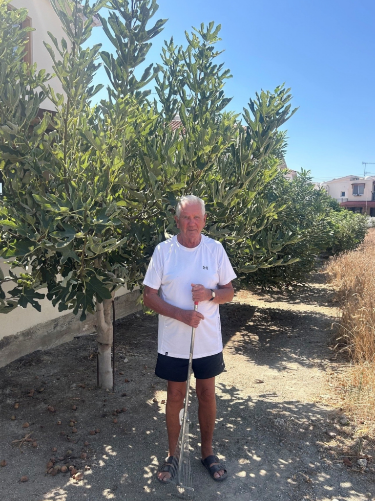 Michael standing outside in Cyprus holding a garden rake in front of tall, leafy fig trees on a sunny day.