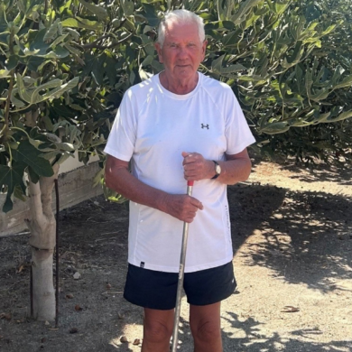 Michael standing outside in Cyprus holding a garden rake in front of tall, leafy fig trees on a sunny day.