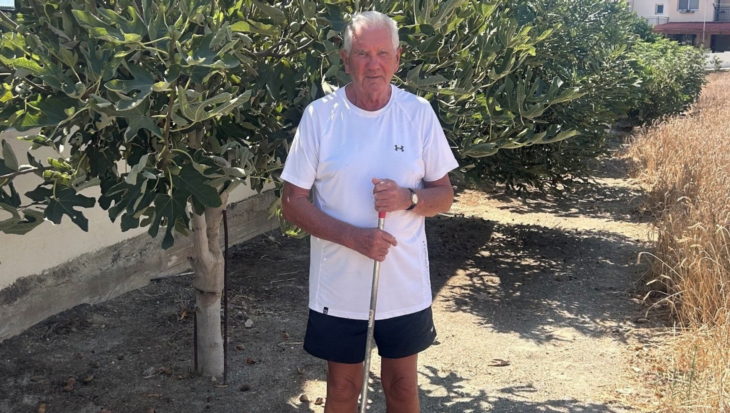 Michael standing outside in Cyprus holding a garden rake in front of tall, leafy fig trees on a sunny day.