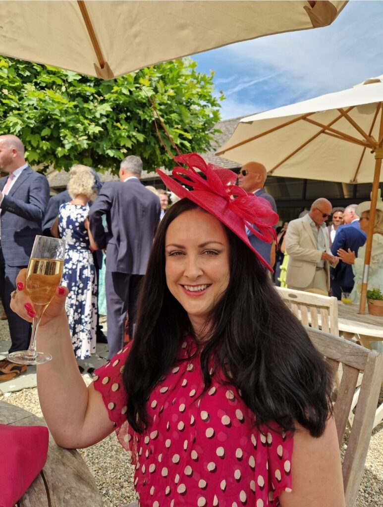 A woman with long dark hair, smiles at the camera and raises a glass of prosecco. She is wearing a red polka dot dress and a red fascinator.