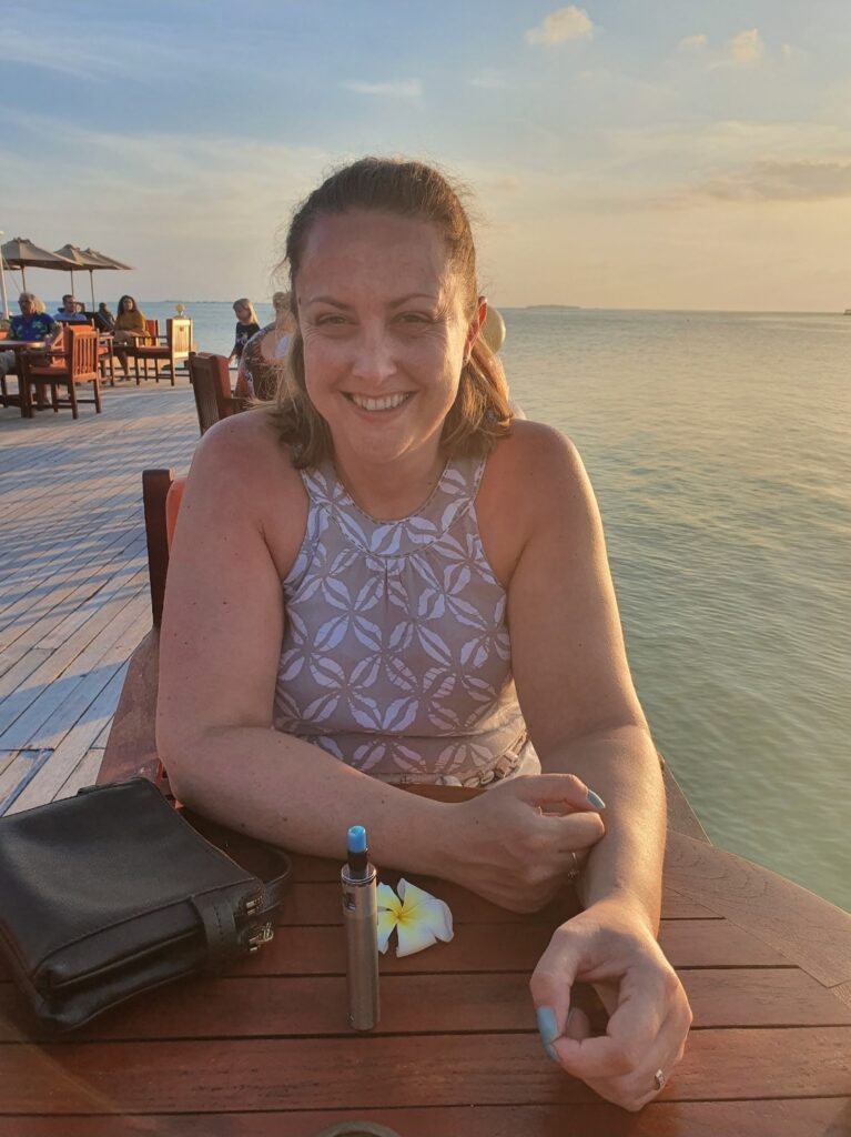 A woman with shoulder length brown hair, sat at a wooden table with a view of the sea and a beach in the background.