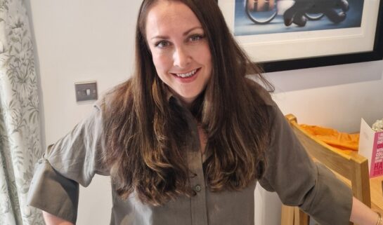 A woman with long dark hair stands in a dining room smiling at the camera. One hand is on her hip, the other resting on a chair.