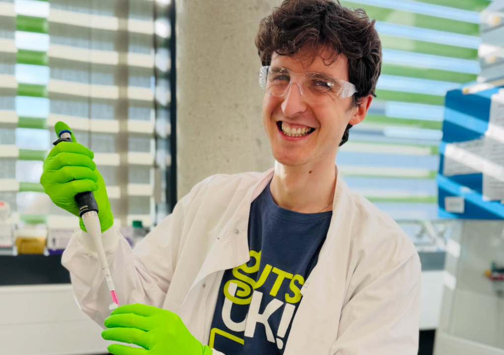 A photograph of a researcher in a lab, wearing a white lab coat and green gloves.