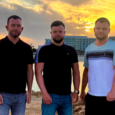 Three men stand with their arms by their sides looking at the camera. Danny is on the left, with his two brother middle and right. The backdrop is a beach and the sea on a holiday.