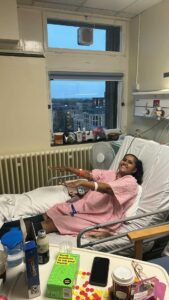 Elizabeth is lying down in a hospital bed wearing a hospital gown and is smiling to the camera. She has both of her arms up in front of her.