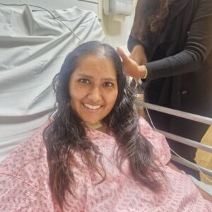 Elizabeth is wearing a pink gown whilst laying down in a hospital bed during her hospital stay. There is also a hand of someone unseen in the picture touching her head.